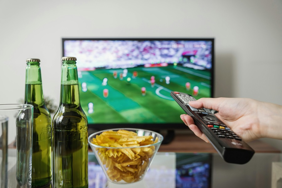 Super Bowl Appetizers a Person Pointing a Remote at a TV with a Game On and Beer Bottles in View with a Bowl of Chips