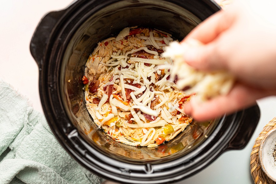 Funny Slow Cooker Memes Overhead View of a Crockpot with Chicken Cooking Inside and a Person's Hand Sprinkling Shredded Cheese Into the Pot