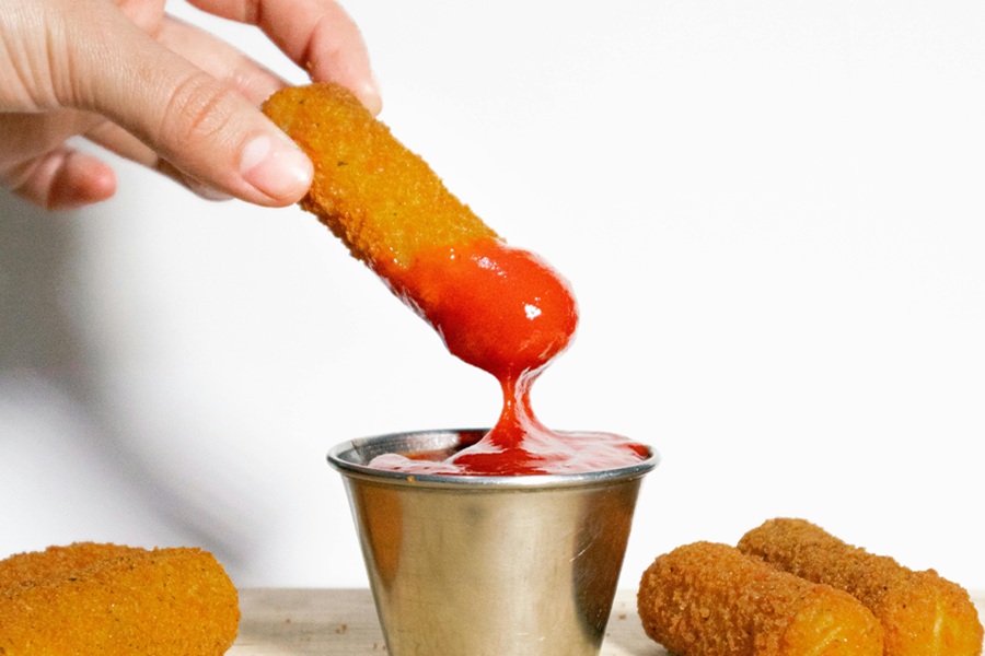 Cheap Air Fryer Recipes Close Up of a Person's Hand Holding a Mozzarella Stick Dipped in Marinara Sauce Hovering Above a Ramekin of Marinara Sauce