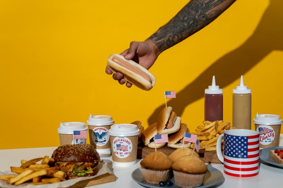 Easy Patriotic Food Appetizers for a Party Close Up of a Table with Hot Dogs and Hamburgers and Coffee Cups Covered in American Flags with a Person's Hand Grabbing a Hot Dog in a Bun