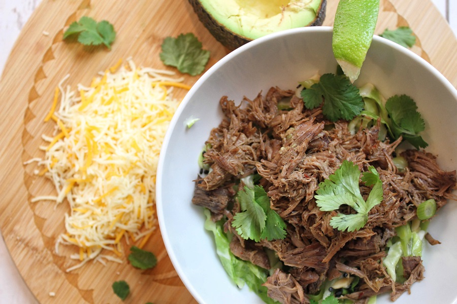 Easy Crockpot Beef Barbacoa in a White Bowl on a Placemat with Shredded Cheese, Avocado, and Cilantro