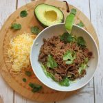 Easy Crockpot Beef Barbacoa a White Bowl Filled with Barbacoa on a Circle Placemat with Half an Avocado, Shredded Cheese, and Cilantro