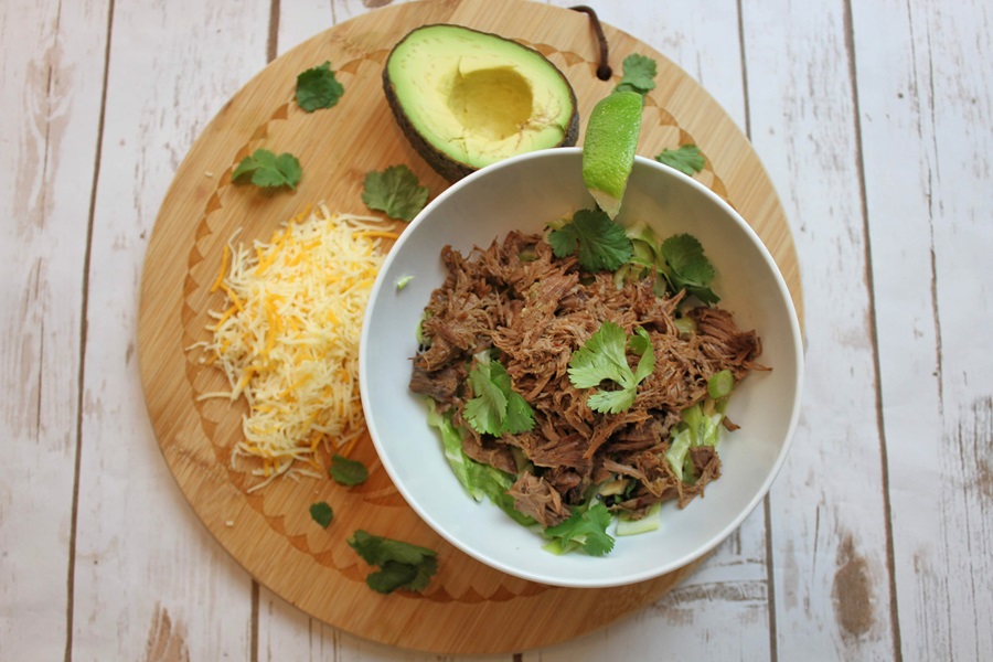 Easy Crockpot Beef Barbacoa a White Bowl Filled with Barbacoa on a Circle Placemat with Half an Avocado, Shredded Cheese, and Cilantro