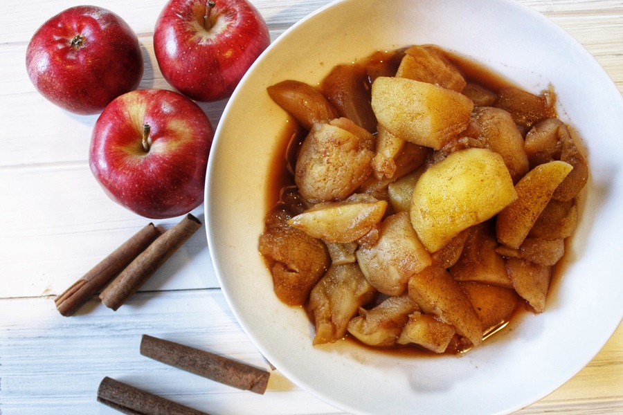 Faster than Stovetop Instant Pot Cinnamon Apples Recipe Overhead View of a White Bowl Filled with Cinnamon Apples Next to Red Apples and Cinnamon Sticks