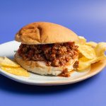 Crockpot Sloppy Joes Recipe a Sloppy Joe on a Plate with Potato Chips on a Lavender Background