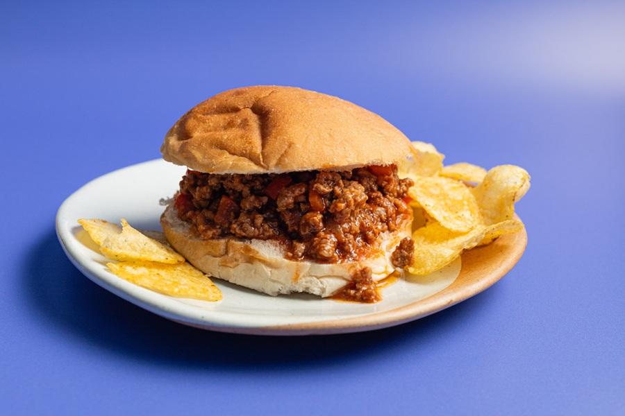 Crockpot Sloppy Joes Recipe a Sloppy Joe on a Plate with Potato Chips on a Lavender Background